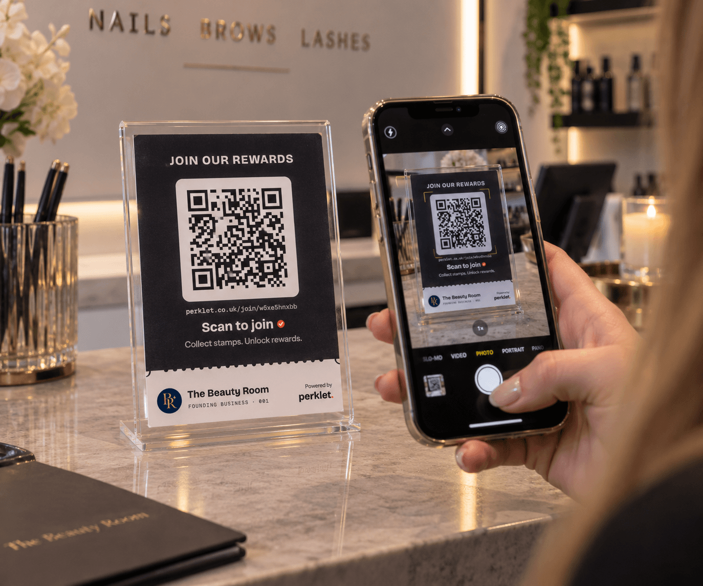A customer at The Beauty Room's till scans the Perklet counter card with their phone camera. The card in its acrylic holder reads “Join our rewards”, with a QR code and “Scan to join”.